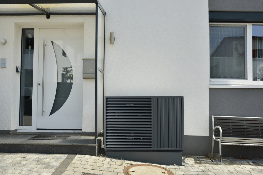 A black heat pump in front of the white façade of a home.